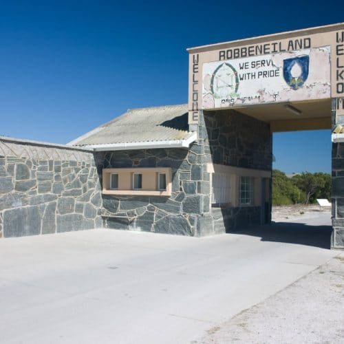 The image captures the entrance to Robben Island, featuring a stone building and an archway labeled "Robben Island Welkom." The sign proudly displays emblems and the slogan "We Serve With Pride" against a clear blue sky, reminiscent of Kruger Tours in Cape Town.