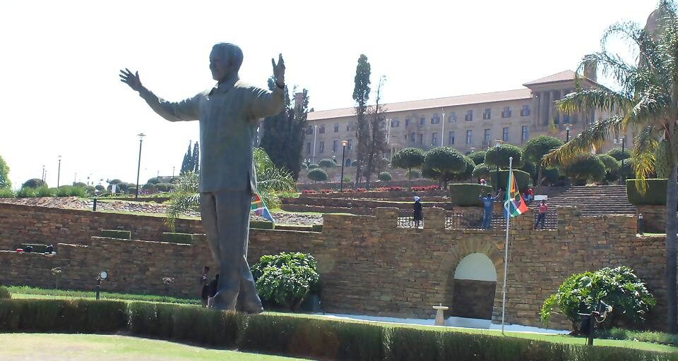 A large bronze statue stands with outstretched arms in a landscaped garden in Pretoria City. The backdrop features the Union Buildings, framed by neatly trimmed shrubs and tall trees. A flagpole bearing the South African flag is prominently visible in the foreground.