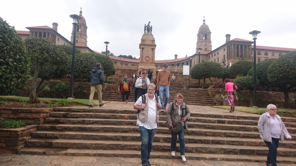 People strolling down stone steps in front of a historic building with tall towers and a central statue in Pretoria City. The picturesque area is surrounded by manicured gardens under cloudy skies, creating an enchanting atmosphere.