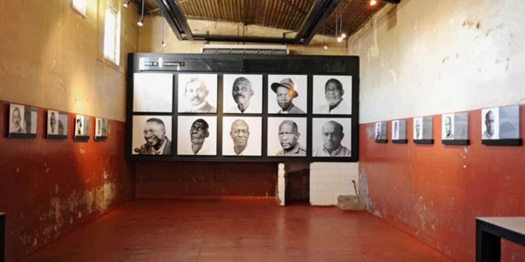 A dimly lit room with framed black-and-white portraits on the walls, featuring several individuals, reminiscent of Constitutional Hill. The room's red floor and rustic, textured walls create a historic atmosphere, while ceiling lights highlight the gallery-style display.