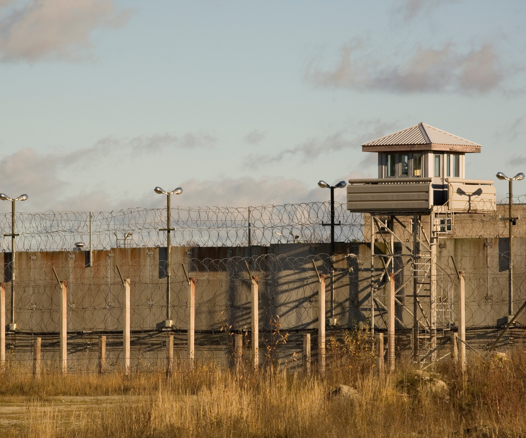 A prison yard with a tall perimeter fence topped with barbed wire mirrors the isolation found far from a full day Cape Winelands tour. A guard tower stationed at the corner overlooks the grassy ground, under a partly cloudy sky.