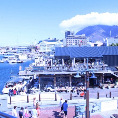 A sunny waterfront scene featuring boats docked at a marina, a boardwalk with people strolling, and a two-story building with outdoor seating. In the background, theres a mountain partially covered by clouds under a clear blue sky.