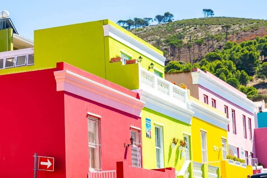 Brightly colored houses in shades of pink, yellow, and green line a street, set against a backdrop of lush greenery and the majestic Table Mountain. This vibrant and cheerful neighborhood scene captures the essence of a day out exploring Cape Town with views reminiscent of a Robben Island tour.