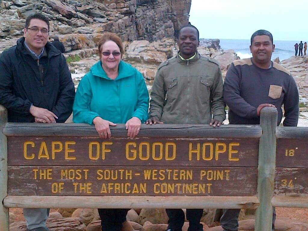 Four people stand together at the Cape of Good Hope sign, which marks the most south-western point of the African continent. The rocky coastline and ocean are visible in the background.