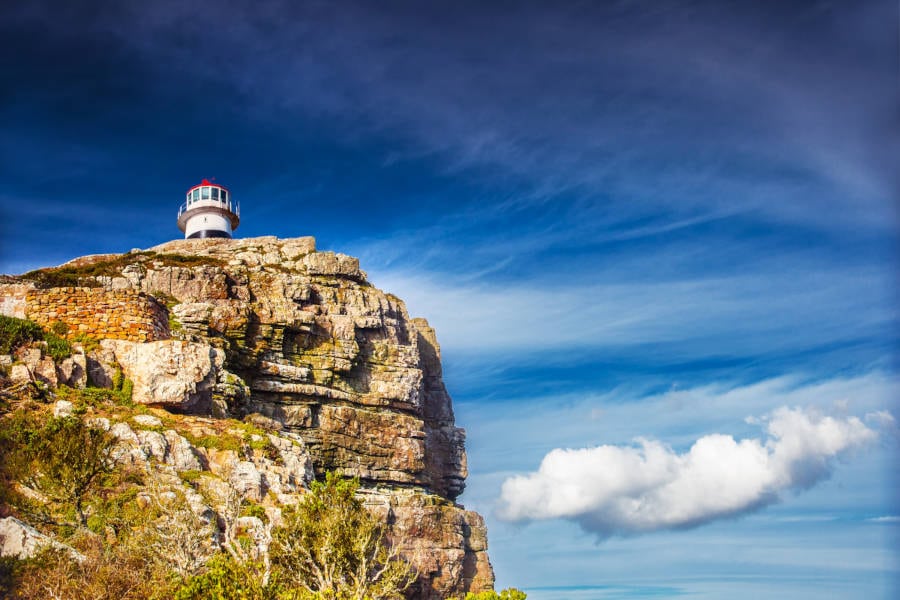 A lighthouse perched atop a rugged cliff under a vivid blue sky with wisps of cloud, much like the scenic views on a Cape Peninsula tour. The landscape is dotted with sparse greenery, conveying solitude and natural beauty.