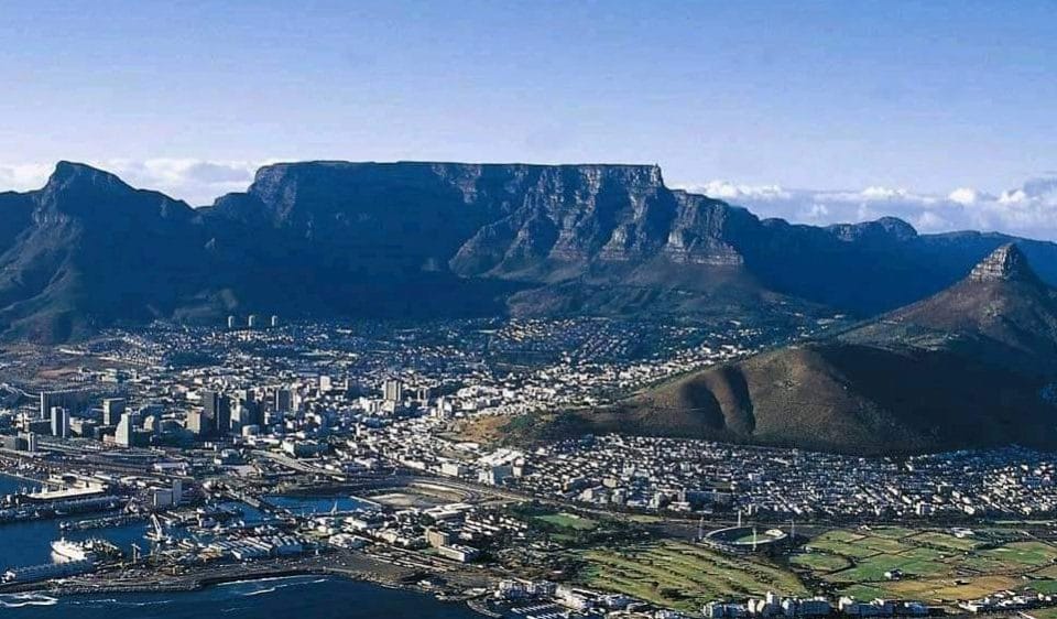 Aerial view of a coastal city where 10 Things to do in Cape Town come to life, with the prominent Table Mountain majestically in the background. The city boasts numerous waterfront buildings, green areas, and smaller hills. The ocean graces the foreground, completing this vibrant tableau.