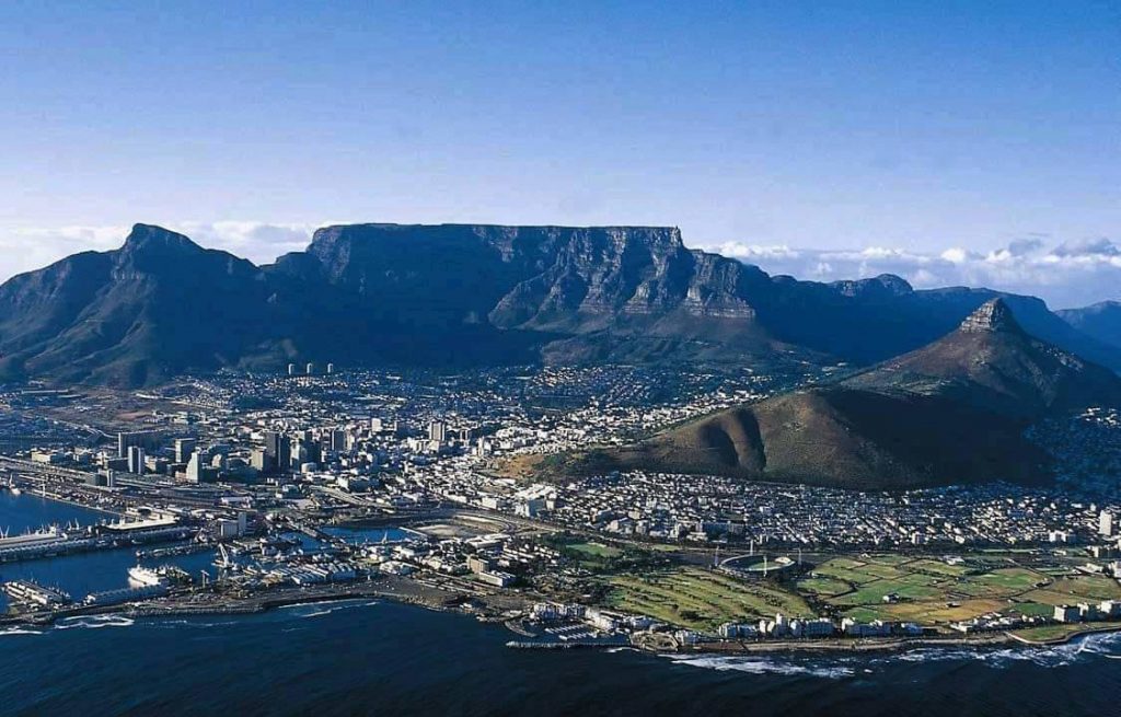 Aerial view of Cape Town, South Africa, with Table Mountain as a prominent backdrop. The cityscape includes modern buildings, and the coastline features a harbor with docks. The foreground shows a grassy area and roads, leading to the ocean.