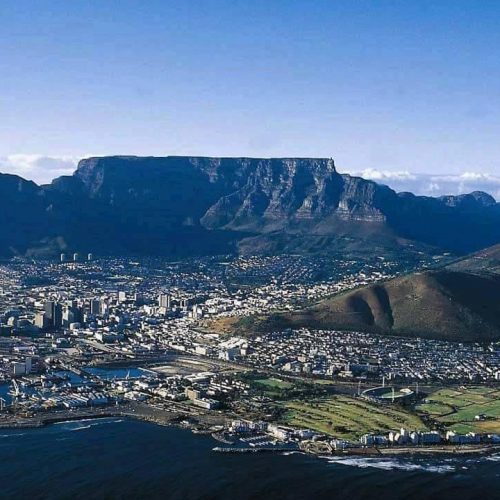 Aerial view of Cape Town, South Africa, with Table Mountain as a prominent backdrop. The cityscape includes modern buildings, and the coastline features a harbor with docks. The foreground shows a grassy area and roads, leading to the ocean.