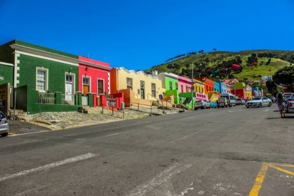 Colorful row of houses on a sloped street, their vibrant green, red, yellow, and pink facades echoing the lively vibe of a Robben Island and Table Mountain tour. Several cars parked along the street with a lush green hill in the background under a bright blue sky.