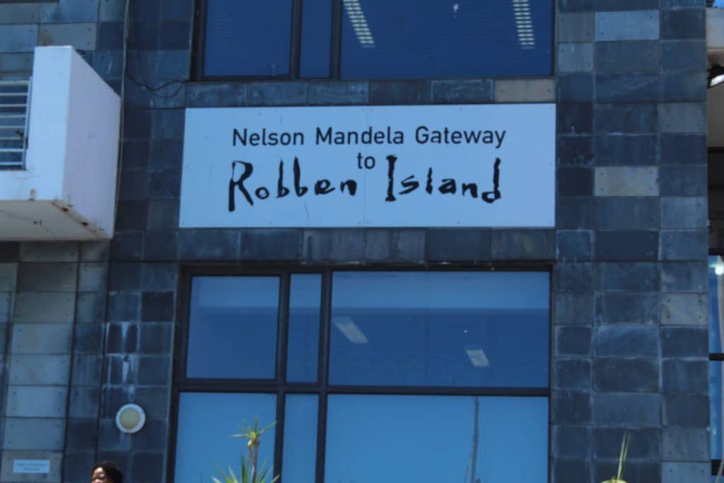 Exterior view of a building with a sign reading Nelson Mandela Gateway to Robben Island. The structure features large windows and gray stone tiles.