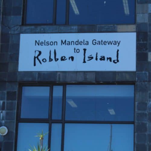 Exterior view of a building with a sign reading Nelson Mandela Gateway to Robben Island. The structure features large windows and gray stone tiles.