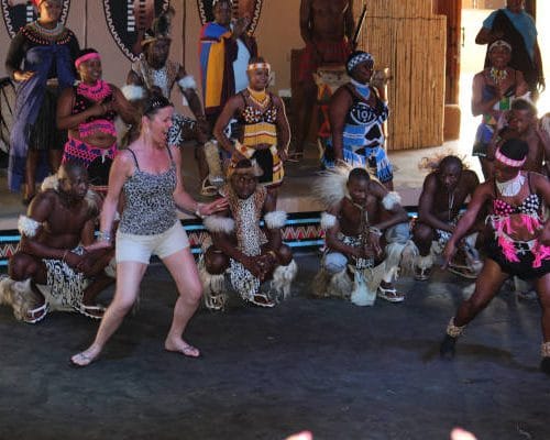 A group of people in traditional attire performs a cultural dance, with a woman in casual clothes joining and mirroring their movements. The indoor setting, reminiscent of Johannesburg's rich heritage, features tribal shields and drums—a perfect glimpse into what 15 days in Kruger might offer.