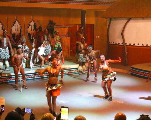 In Johannesburg, during a vibrant 15-day festival, a group of dancers in traditional attire performs energetically on stage. Drums and shields provide a rhythmic backdrop as the audience records the lively display with their phones, surrounded by ethnic decor that mirrors Kruger's cultural richness.