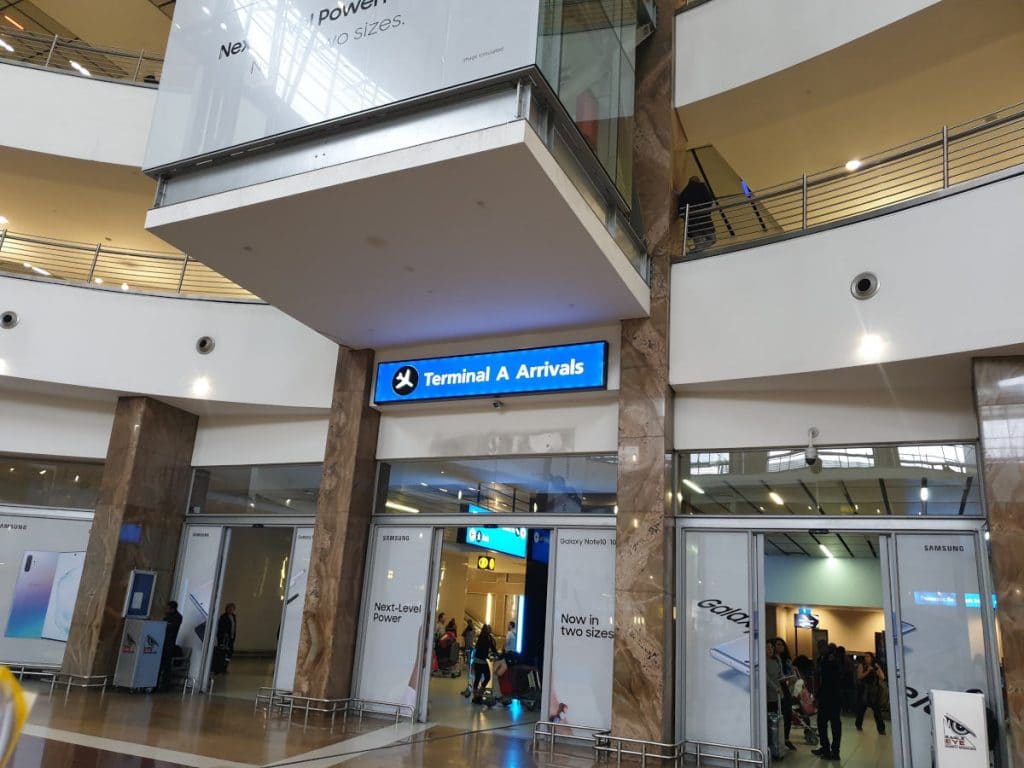 An airport terminal with a sign reading Terminal A Arrivals. People are gathered near the entrance. The interior features large glass panels and advertisements on the walls.