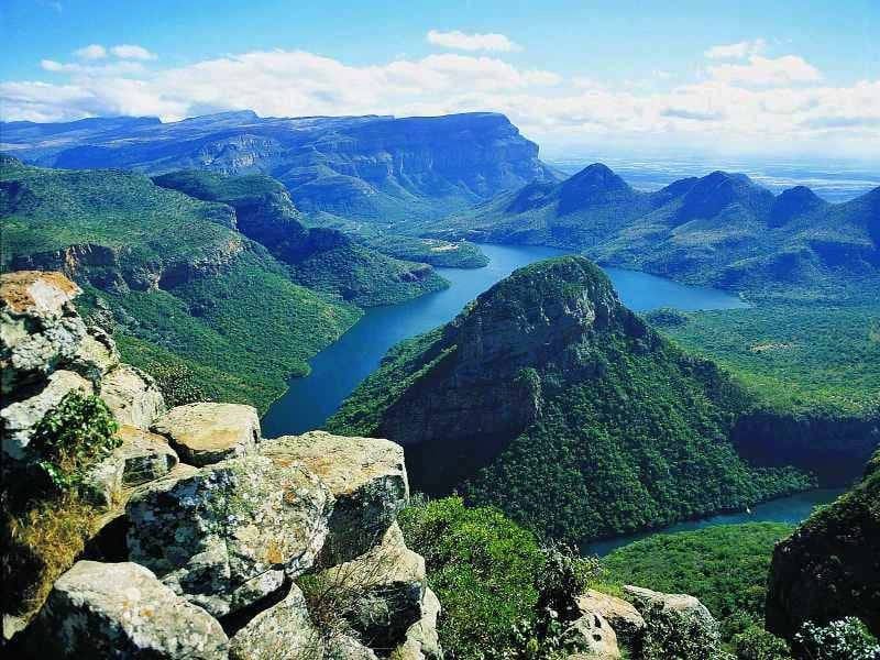 A breathtaking view of a lush, green canyon with a winding river below. Rocky cliffs frame the foreground, while distant mountains rise under a bright blue sky scattered with fluffy clouds.