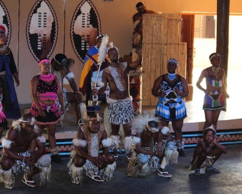 A group of people in traditional attire perform a cultural dance indoors, reminiscent of Johannesburg's rich heritage. They wear vibrant, patterned costumes with feathers and beads. Behind them are decorative shields and a thatched wall, enhancing the cultural ambiance like a scene from 15 Days in Kruger.