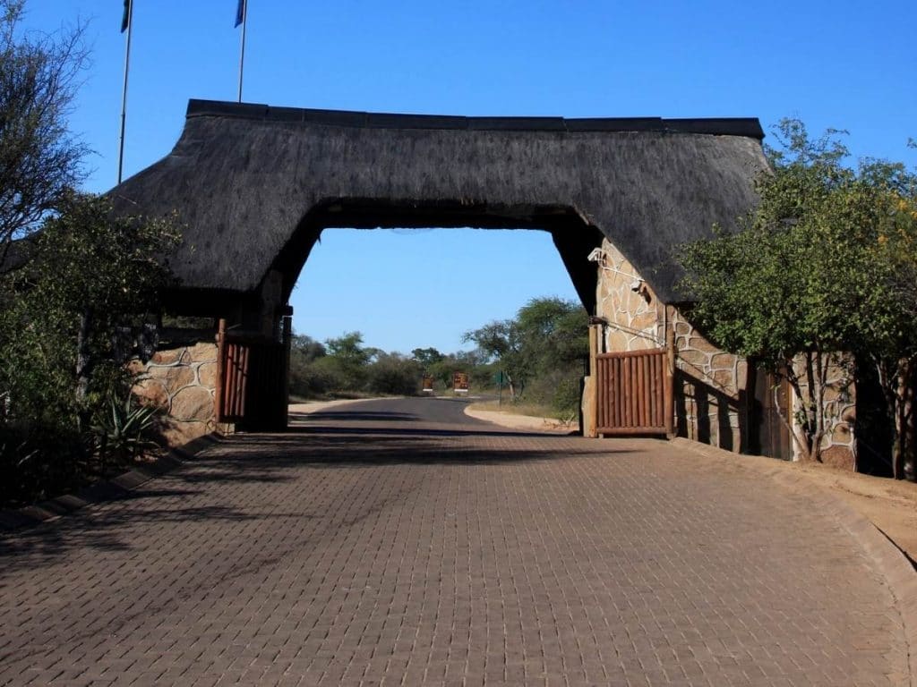 A wide gate with a thatched roof spans across a paved road, flanked by stone walls and trees. The road leads into a stunning natural reserve, reminiscent of the scenic landscapes one might explore on a 6-day Kruger and Johannesburg adventure under a clear blue sky.