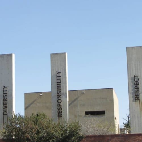 A series of tall concrete pillars with the words Democracy, Reconciliation, Diversity, Responsibility, Respect, and Freedom etched on them stand proudly beneath Johannesburg's clear blue sky. A building and trees quietly frame this striking scene reminiscent of a memorable 5-day journey from Cape Town.