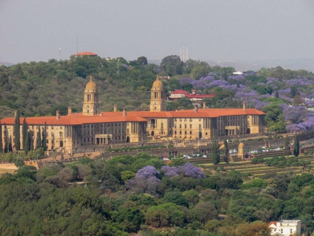 A large historic building with red roofs and twin domes sits among lush greenery and blooming jacaranda trees under a clear sky, much like the serene landscapes you'll encounter on a 6-day journey through Kruger and Johannesburg, set against a backdrop of rolling hills.