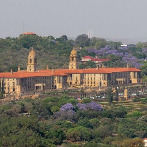 A large historic building with red roofs and twin domes sits among lush greenery and blooming jacaranda trees under a clear sky, much like the serene landscapes you'll encounter on a 6-day journey through Kruger and Johannesburg, set against a backdrop of rolling hills.