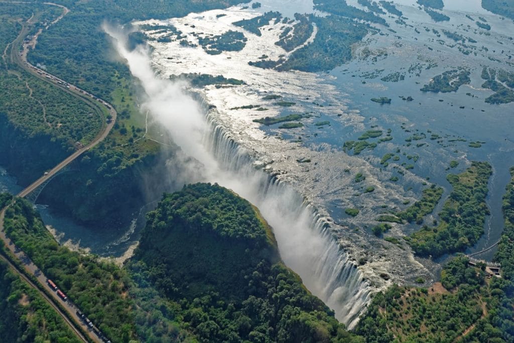 Aerial view of a large waterfall cascading over a cliff surrounded by lush greenery and mist, with a river and railway track visible to the side.