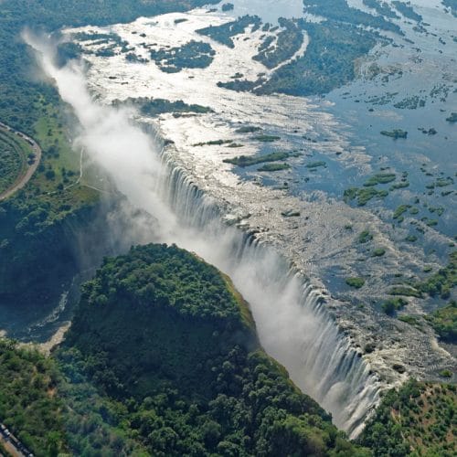 Aerial view of a large waterfall cascading over a cliff surrounded by lush greenery and mist, with a river and railway track visible to the side.