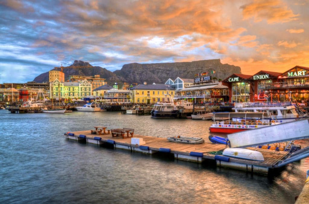 A vibrant harbor scene at sunset with boats docked at a wooden pier. Colorful buildings line the waterfront under a dramatic sky, and a mountain range is visible in the background.