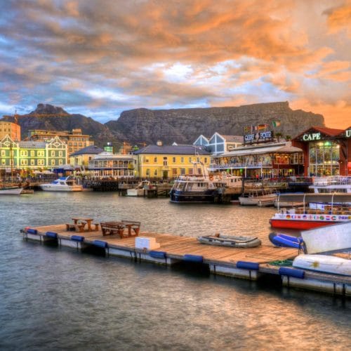 A vibrant harbor scene at sunset with boats docked at a wooden pier. Colorful buildings line the waterfront under a dramatic sky, and a mountain range is visible in the background.