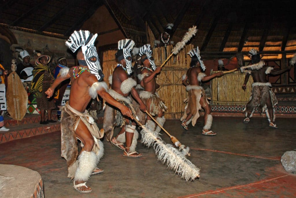 A group of dancers in traditional attire, including feathered headdresses and animal pelts, performs energetically in an indoor setting. The dancers are holding ornate staffs, and the background features cultural decorations.