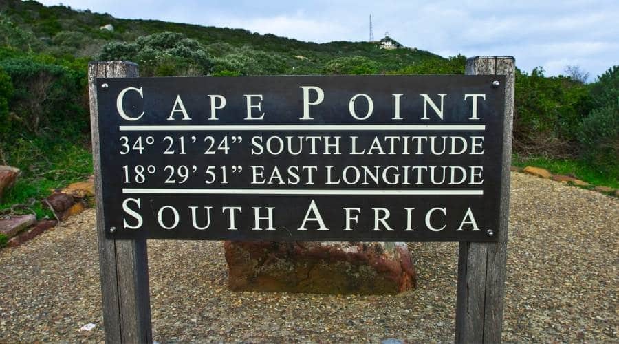 A sign at Cape Point, South Africa, marks coordinates 34° 21' 24" S and 18° 29' 51" E, a highlight of the Best of the Cape Tour. The lush greenery and cloudy sky in the background set a breathtaking scene.