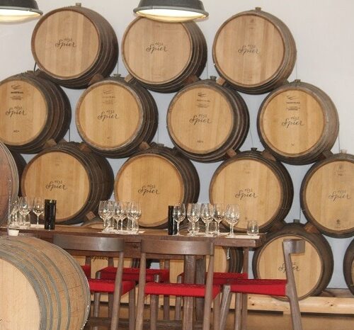 A cozy wine tasting room, often featured in Cape Town Tour Packages, boasts a wooden table adorned with wine glasses and bottles. Behind the table, rows of large wooden barrels are stacked against the wall, crafting a warm and inviting atmosphere.