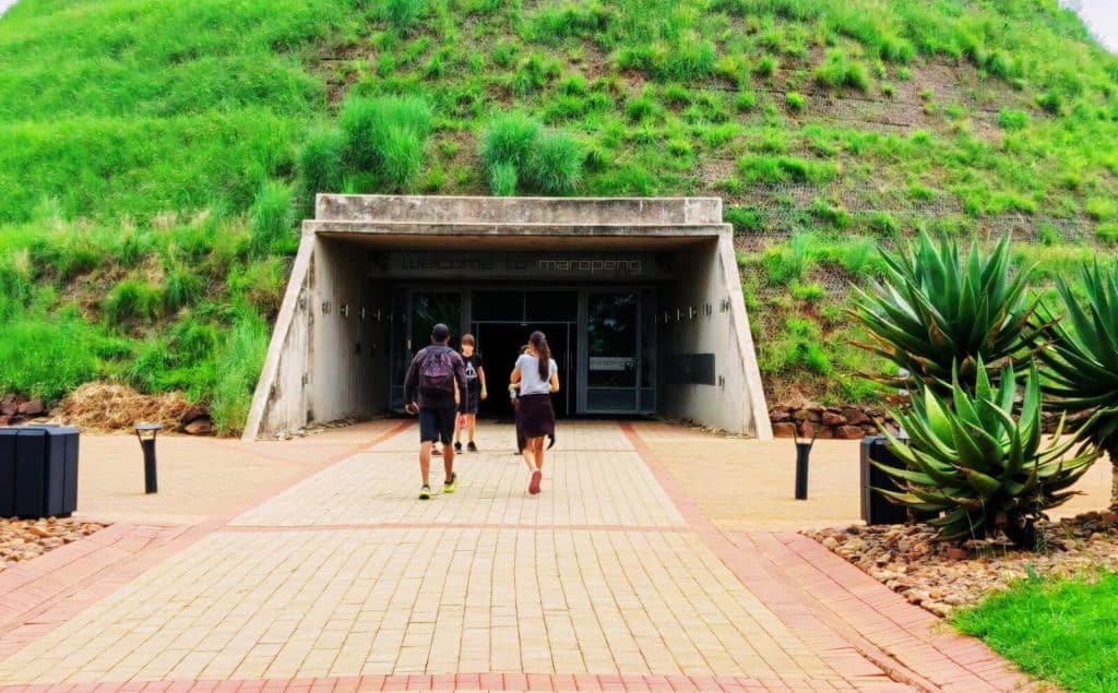 Three people walk towards the entrance of a grass-covered, bunker-like building. The path is paved and surrounded by plants, with the words Welcome to Maropeng visible above the door, inviting them to explore the cradle of humankind tour that awaits inside.