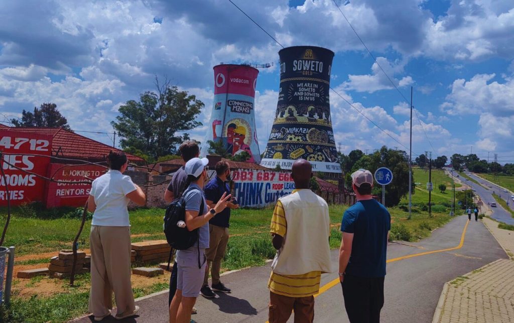 A group of people on a Soweto tour stand on the sidewalk, taking in the vibrant Orlando Towers near the road. The sky is partly cloudy, with banners and signs adding to the lively atmosphere.