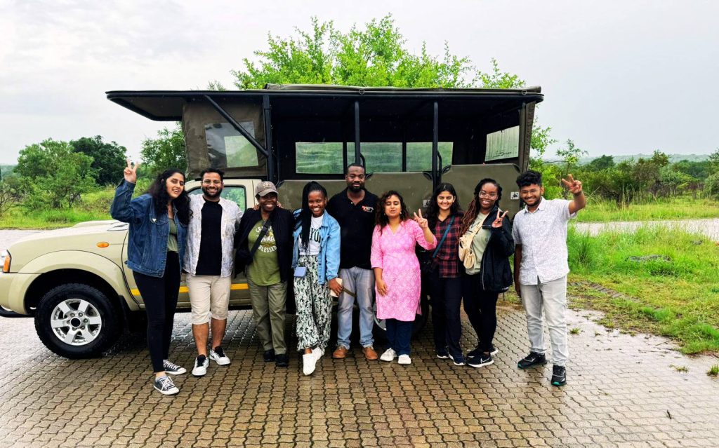 A group of people smiling and posing in front of an open safari vehicle, embarking on their 2-day Kruger safari from Johannesburg. One person is holding up a peace sign amid the lush greenery. The sky is overcast, adding to the anticipation of adventure.