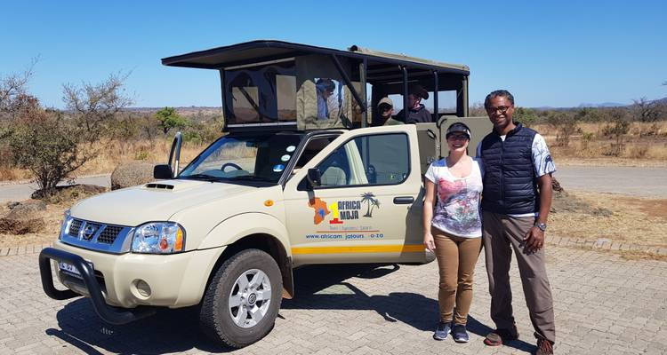 A man and woman stand smiling in front of a safari vehicle with a canopy. The background shows sparse vegetation and a clear sky, suggesting a warm, dry climate.