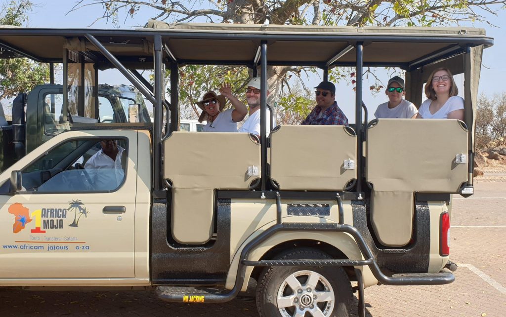 A group of people smiling and waving sit in an open safari vehicle parked on a paved surface during their exciting 2-day Kruger Safari from Johannesburg. The vehicle, proudly displaying the African Moja logo, is surrounded by lush trees in the background.