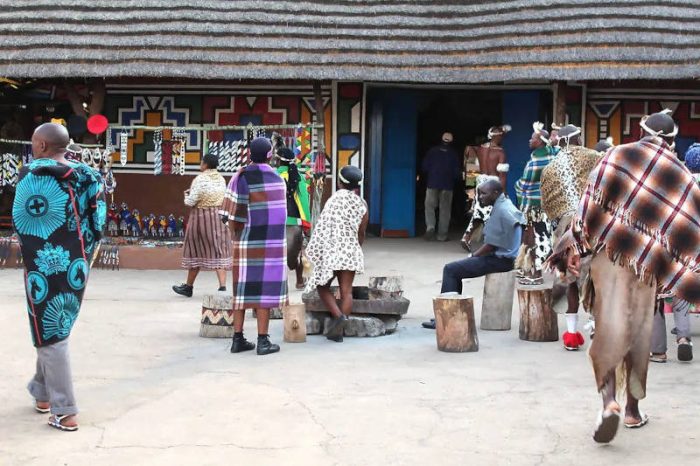 At Lesedi Cultural Village, people in traditional clothing interact, highlighting vibrant patterns amidst a backdrop of geometric buildings. Wooden stools are thoughtfully arranged around a central point, offering an inviting glimpse into this enriching cultural tour.