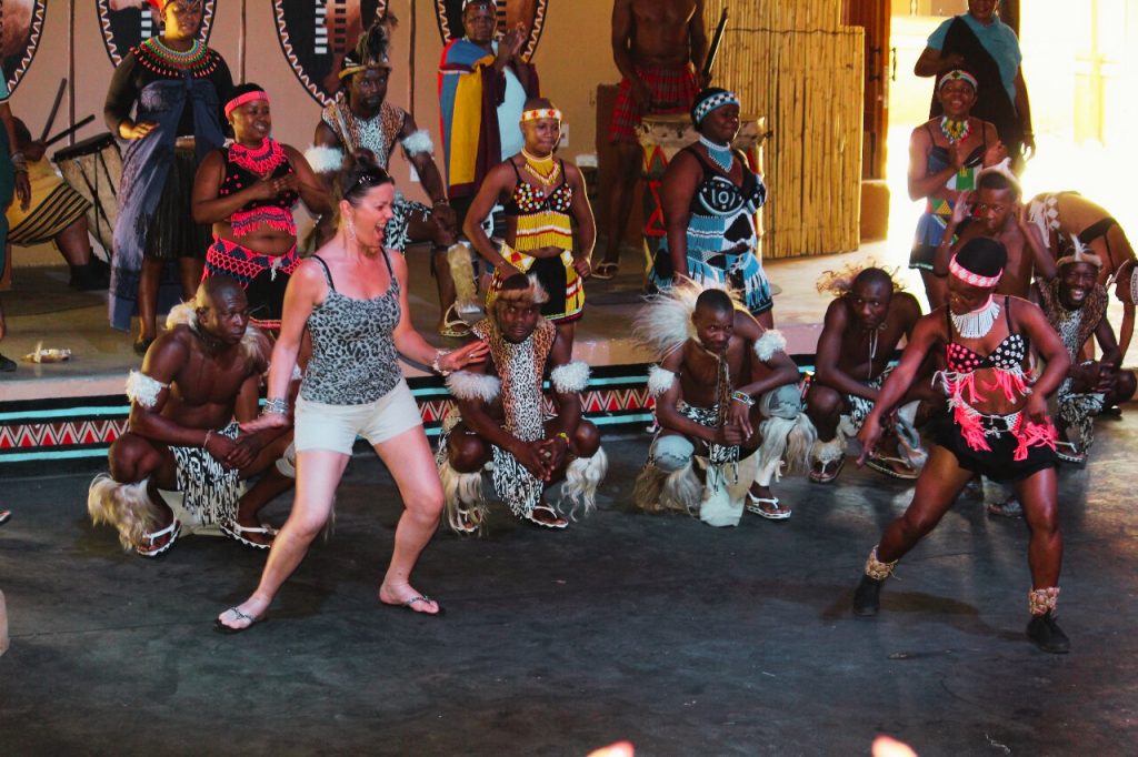 During the Lesedi Cultural Village Tour, a group of people in traditional African attire perform a dance on stage. A woman in casual clothing joins them, mimicking their movements. The dancers wear colorful clothing and adornments, bringing a lively and vibrant energy to the setting.