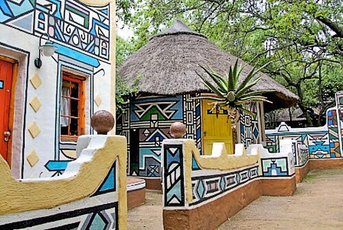 Explore the Lesedi Cultural Village Tour, where colorful buildings boast intricate geometric patterns and bright doors, complemented by a traditional thatched-roof hut. Vibrant artwork adorns the walls and pathways, all nestled amidst green trees in this lively cultural setting.