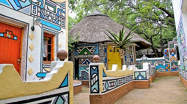 Explore the Lesedi Cultural Village Tour, where colorful buildings boast intricate geometric patterns and bright doors, complemented by a traditional thatched-roof hut. Vibrant artwork adorns the walls and pathways, all nestled amidst green trees in this lively cultural setting.