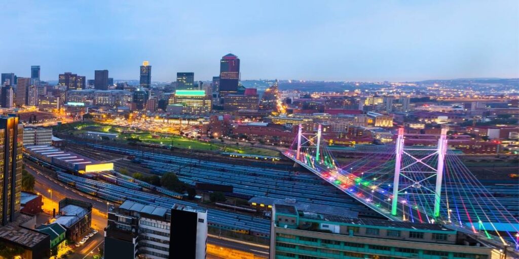 Aerial view of a cityscape at dusk, with illuminated buildings and roads highlighting the vibrant Soweto tour. The colorful, lit-up cable-stayed bridge stands in the foreground, and rail tracks run parallel beneath it, while lights twinkle throughout the urban landscape.