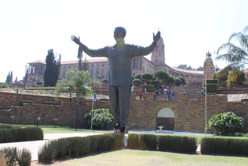 A large statue of a person with outstretched arms welcomes visitors on the Pretoria city tour, standing proudly in front of a historic building with a tower. The sunny, landscaped area features elegant hedges and flagpoles, making it a picturesque stop for tourists.