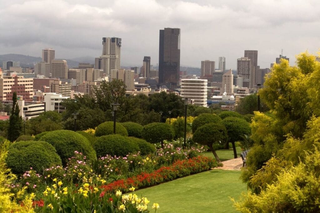 A lush garden with trimmed bushes and colorful flowers overlooks the Pretoria city skyline, featuring tall modern buildings under a cloudy sky—a perfect spot to start your city tour.