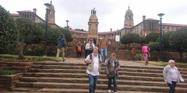 People strolling down stone steps in front of a historic building with tall towers and a central statue in Pretoria City. The picturesque area is surrounded by manicured gardens under cloudy skies, creating an enchanting atmosphere.