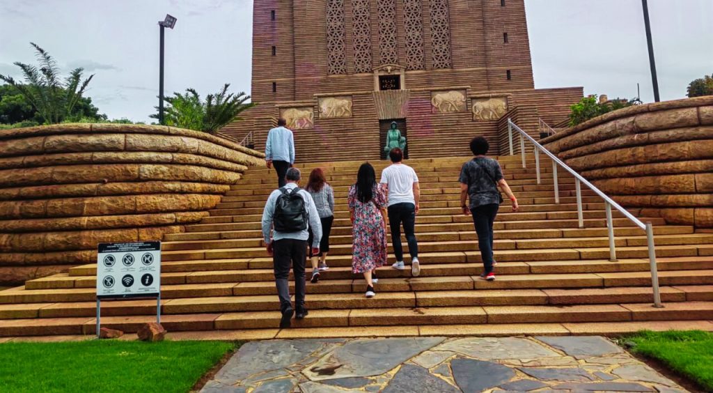 A group of people walking up stone steps towards a large, ornate brown building with a patterned facade, reminiscent of the architecture seen on a Soweto Apartheid Museum tour. The path is flanked by greenery, and a sign with symbols is visible on the left side.