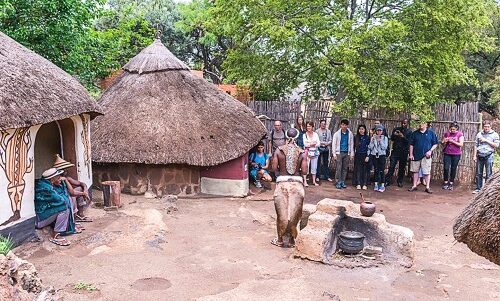 A group of people stand in a semi-circle at the Lesedi Cultural Village Tour, surrounded by traditional thatched-roof huts. Two individuals are seated outside one hut. In the center, a small elephant stands beside a pot on an open fire, with trees and a wooden fence in the background.