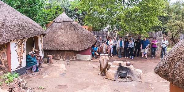 A group of people stand in a semi-circle at the Lesedi Cultural Village Tour, surrounded by traditional thatched-roof huts. Two individuals are seated outside one hut. In the center, a small elephant stands beside a pot on an open fire, with trees and a wooden fence in the background.