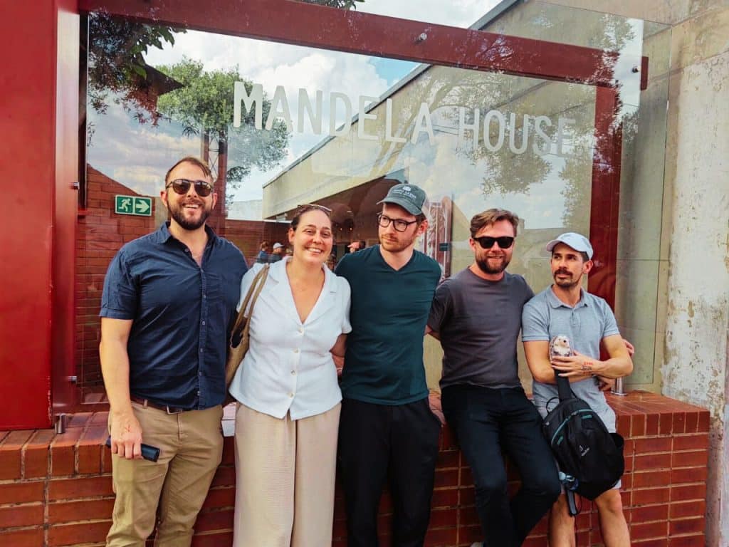 A group of five people, standing close together and smiling, pose for a photo in front of a sign that reads Mandela House. They're capturing memories on their Half Day Soweto Tour outside a building with glass and brick elements. Trees and sky are visible in the reflection.