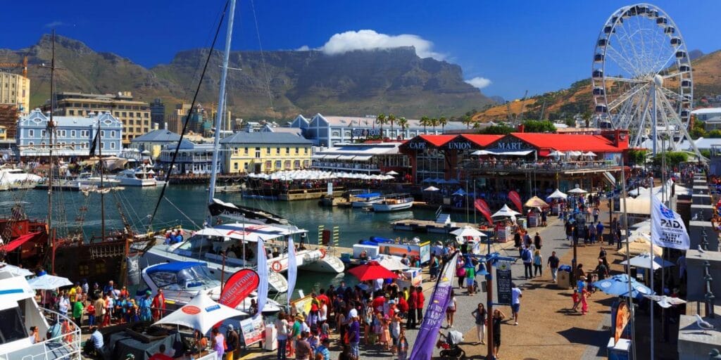 A lively waterfront bustling with tourists exploring Cape Town tour packages, boats docked in a harbor, and a large Ferris wheel. The scene is set against majestic mountains under a clear blue sky, with vibrant shops and people enjoying the day.