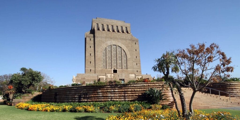 A large, rectangular monument made of stone stands under Pretoria's clear blue sky, surrounded by landscaped gardens and colorful flowers. The structure boasts intricate carvings on its facade and an imposing, monumental design—a must-see on any Pretoria city tour.